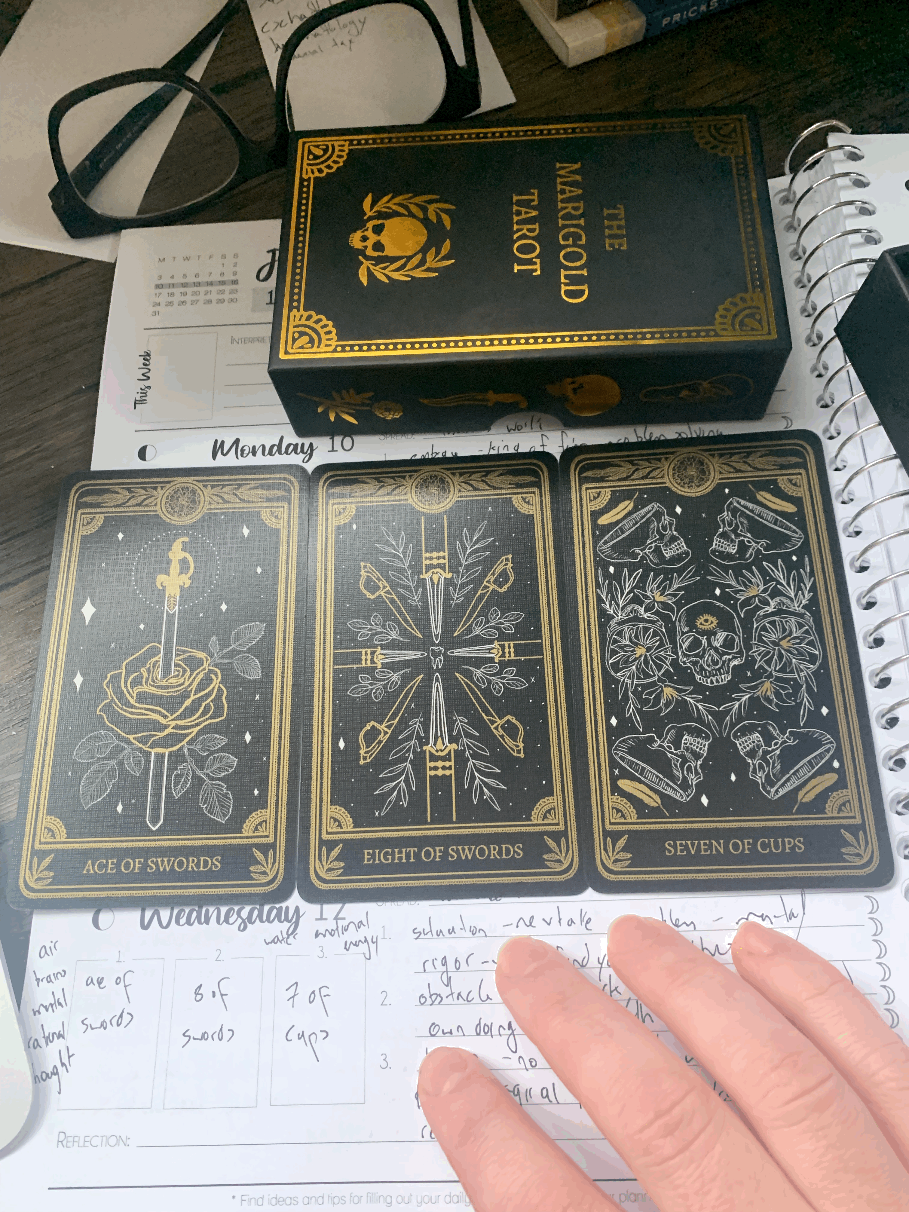 three card black & gold tarot spread: ace of swords, eight of swords, seven of cups on top of a planner notebook