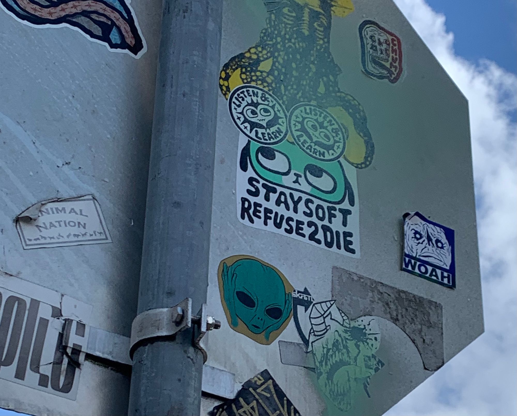 back of a stop sign covered in peeling stickers. one reads: "stay soft refuse2die."