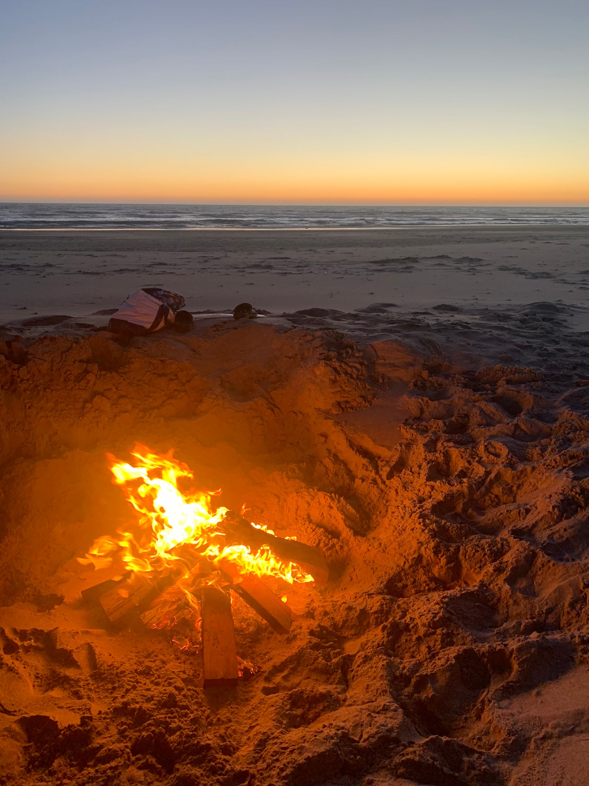 evening, sandy beach, wood fire in foreground, ocean in background, a bag & a pair of shoes perched on the edge of the sandy hole