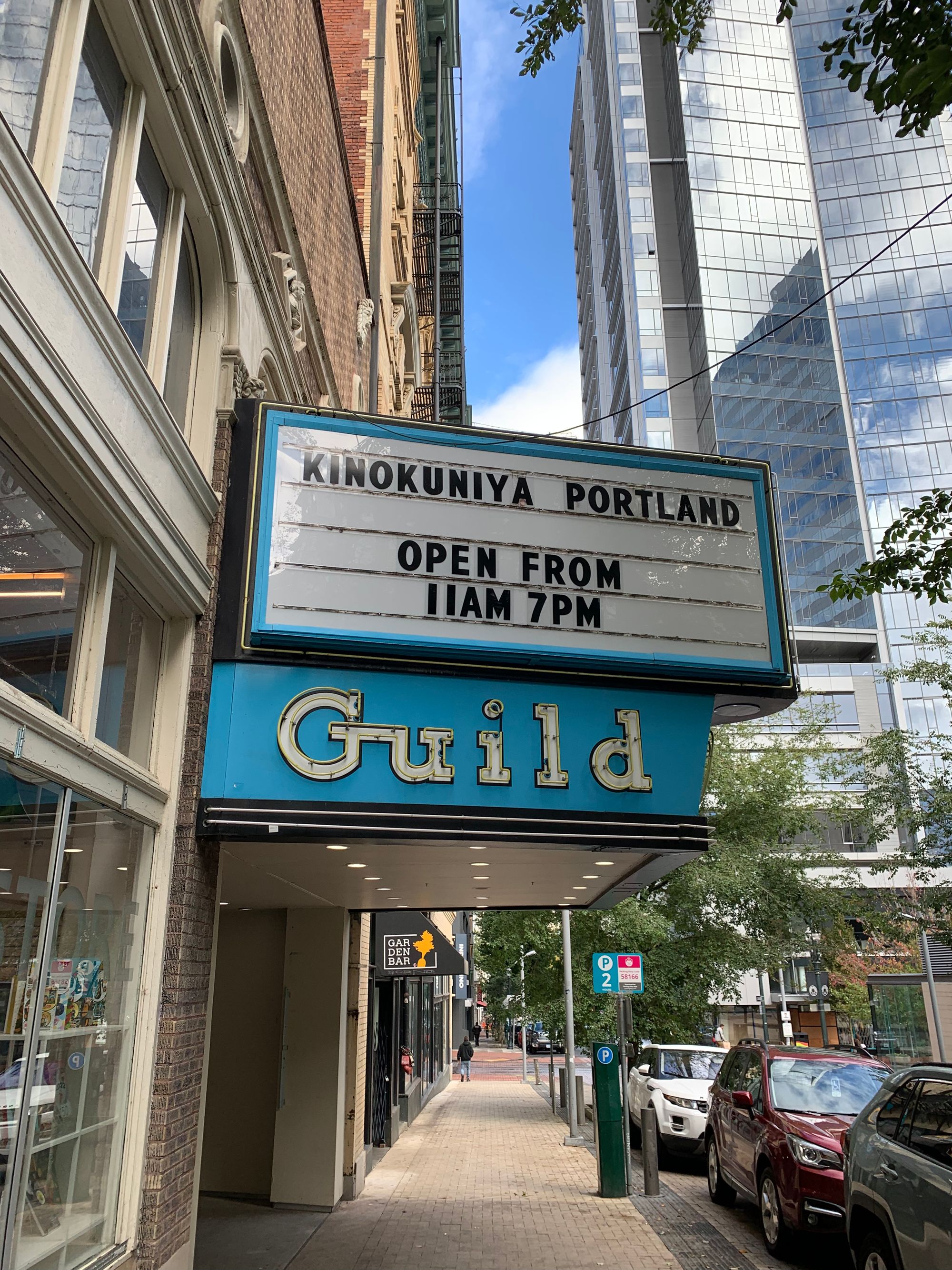 Marquis sign for Kinokuniya Portland, street view of downtown Portland with brick sidewalks, cars & trees, a few people walking in the distance