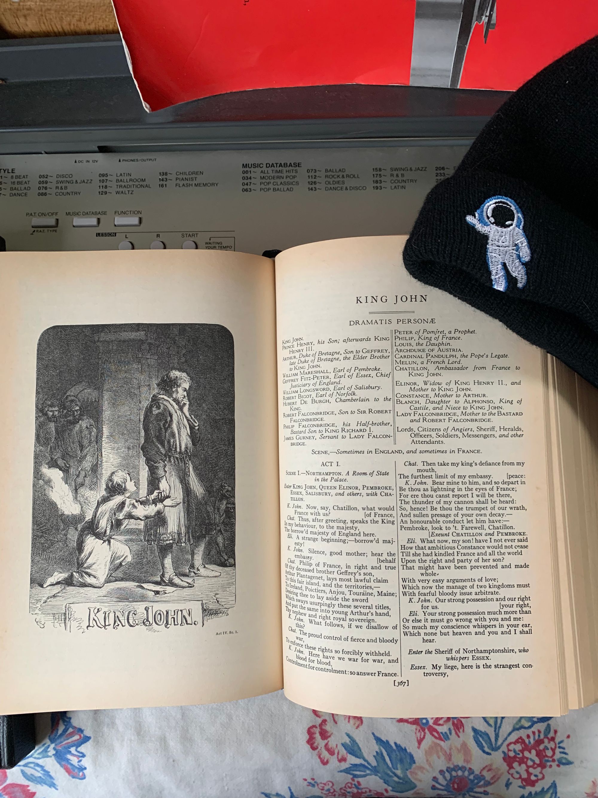 Edition of Shakespeare's plays open to King John, a drawing of King John being implored by Prince Arthur. Book is lying open on a patterned piece of white cloth, on top of a keyboard, with a black hat resting next to it