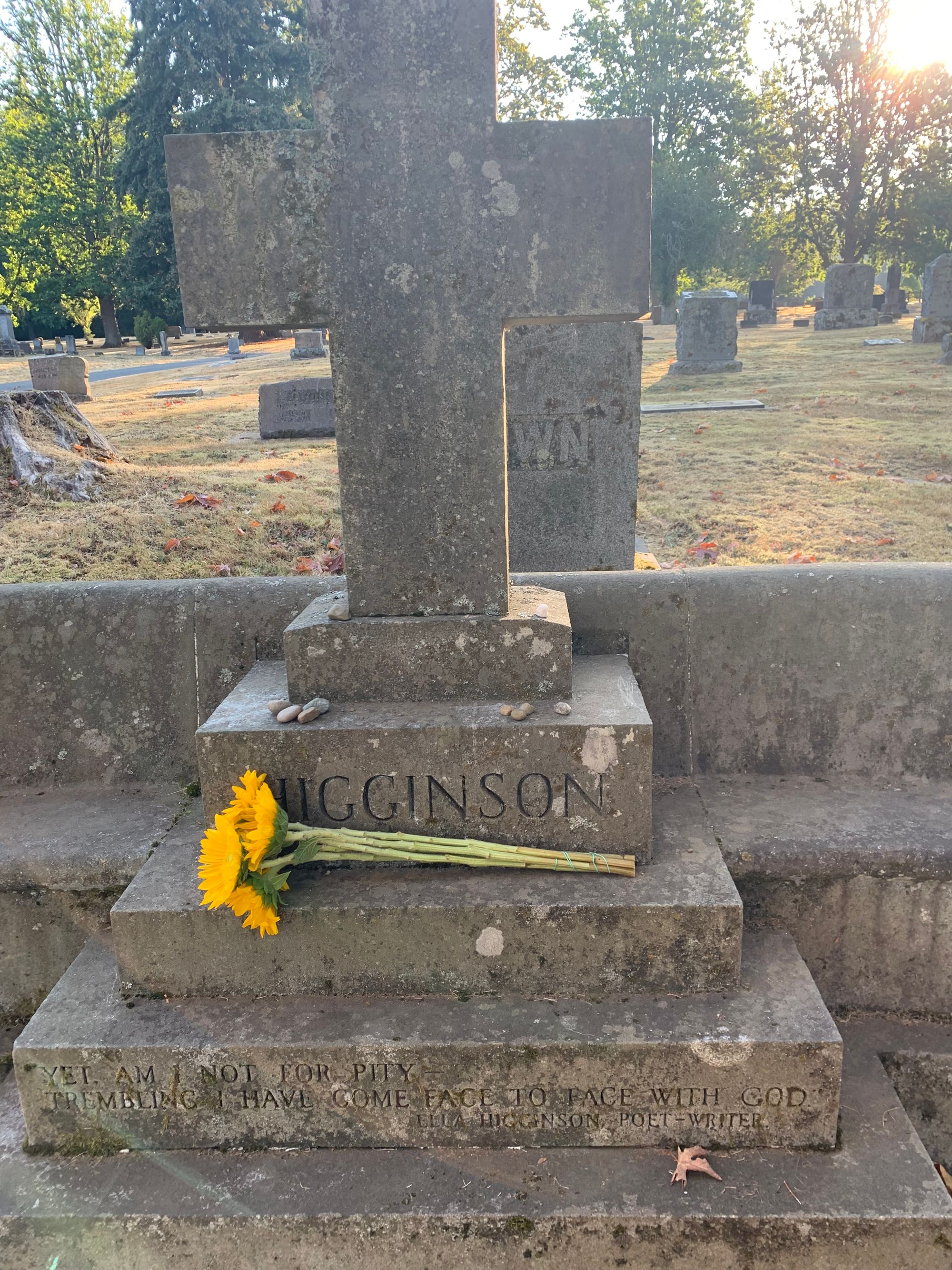 gravestone with large cross, inscription reads: "Yet, am I not for pity/trembling I have come face to face with God-Ella Higginson, poet-writer." Yellow flowers laid on grave.