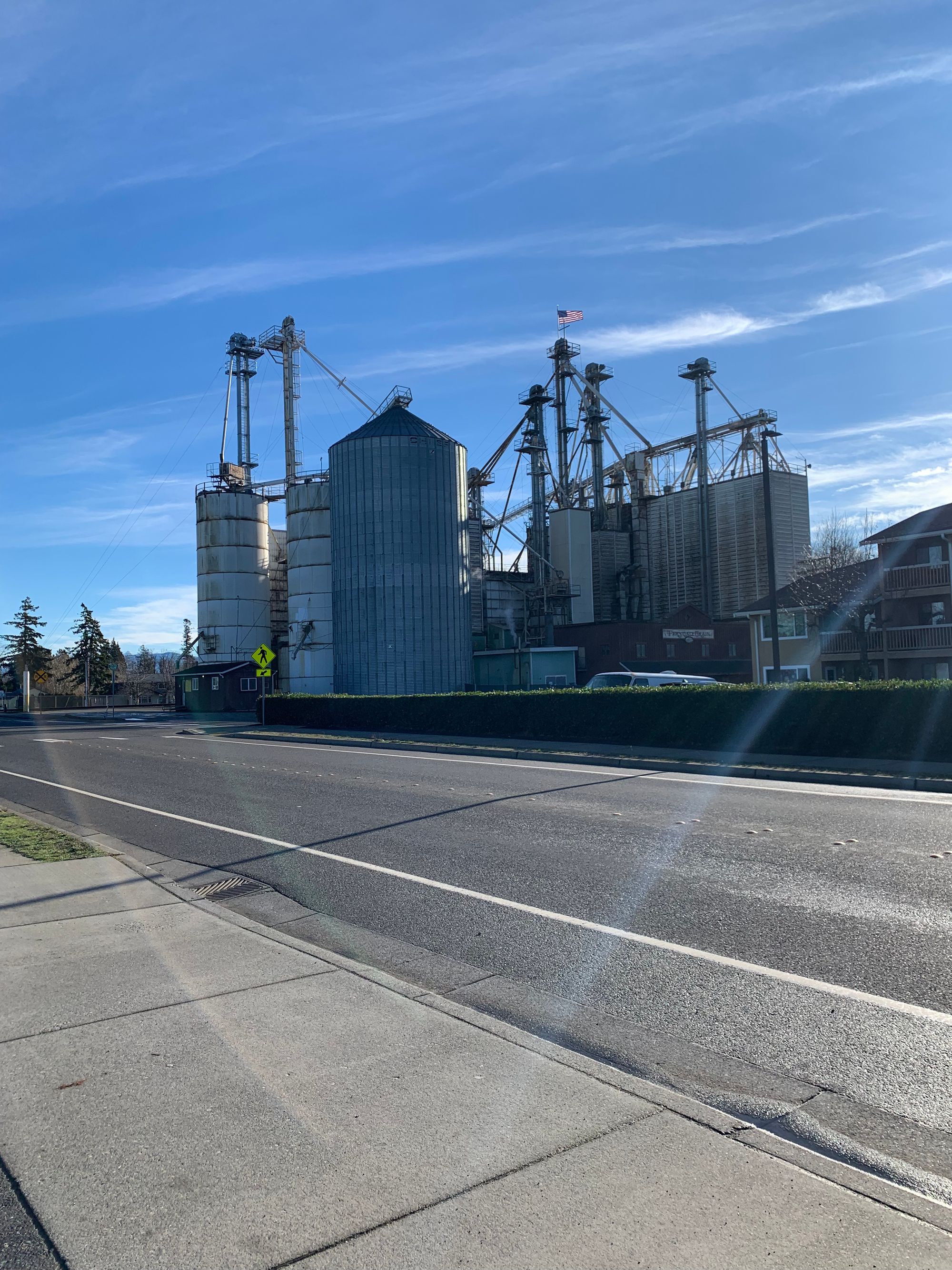 bright blue sky, tall metal towers of a grain processing facility, double lane road & sidewalk in foreground