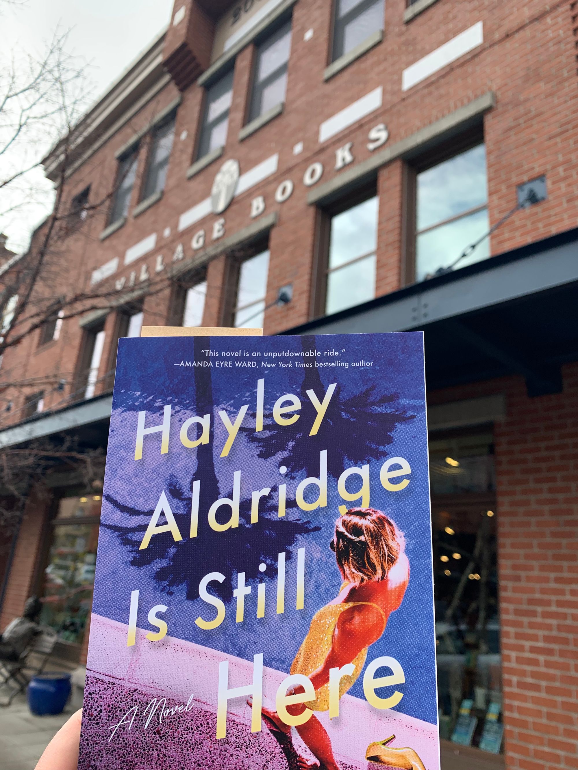 brick bookstore building in background, paperback copy of Hayley Aldridge Is Still Here held up in front of it