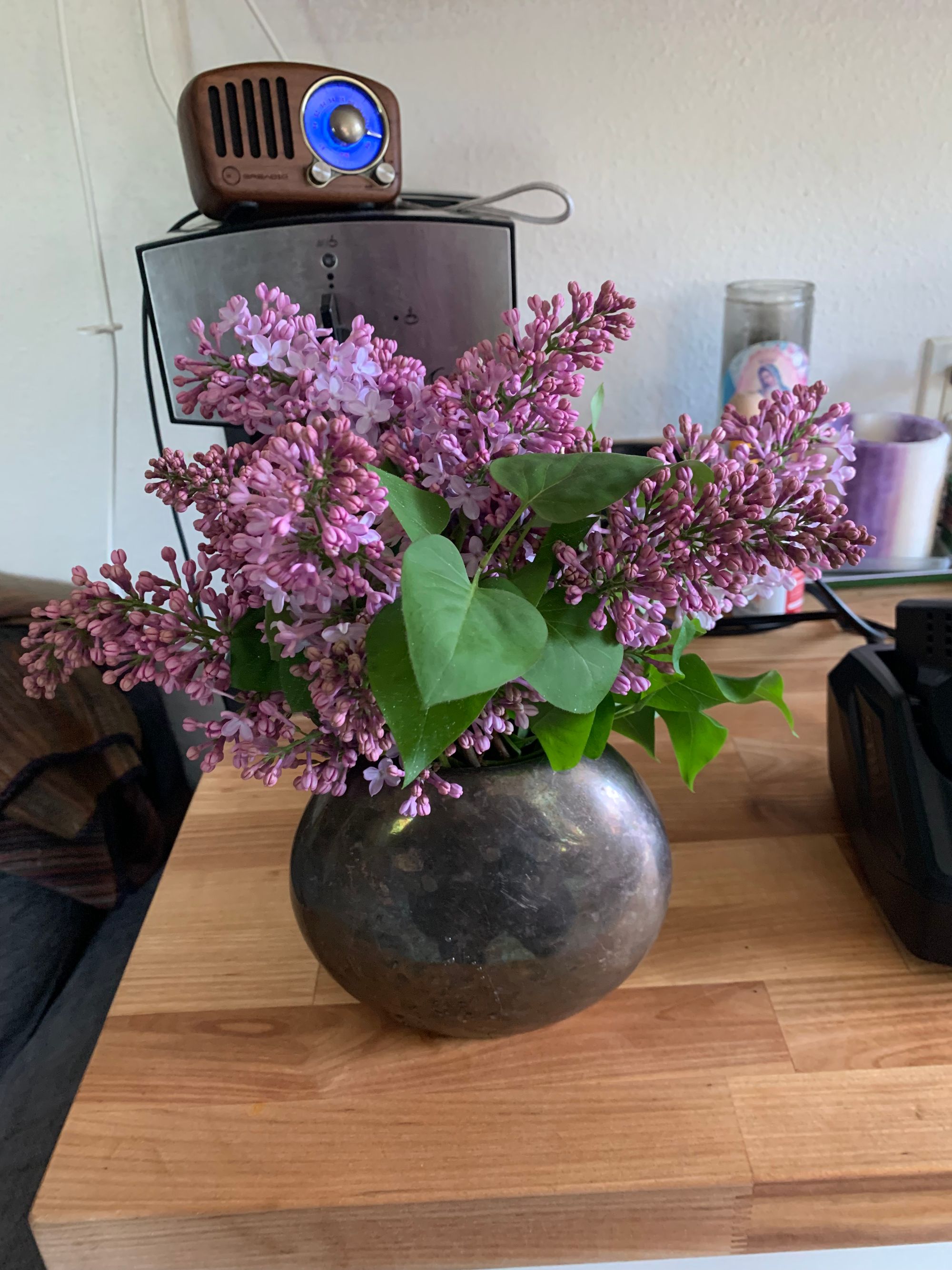 small pewter vase full of light purple lilacs on a butcher block counter top, coffee maker & little radio in the background