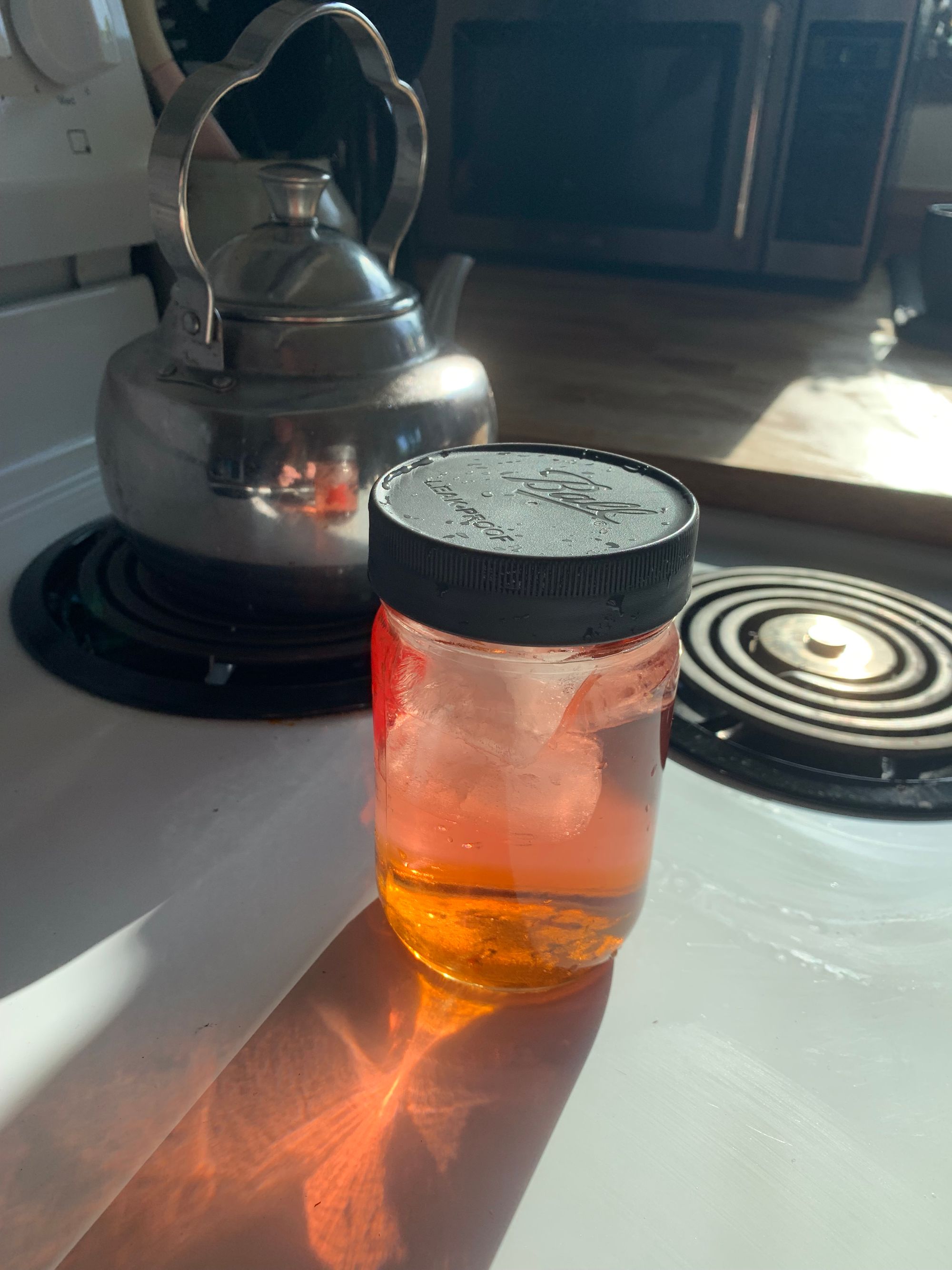 glass jar of pink-brown iced tea with a grey lid sitting on a white stovetop with sun shining through & making a reflection of the jar