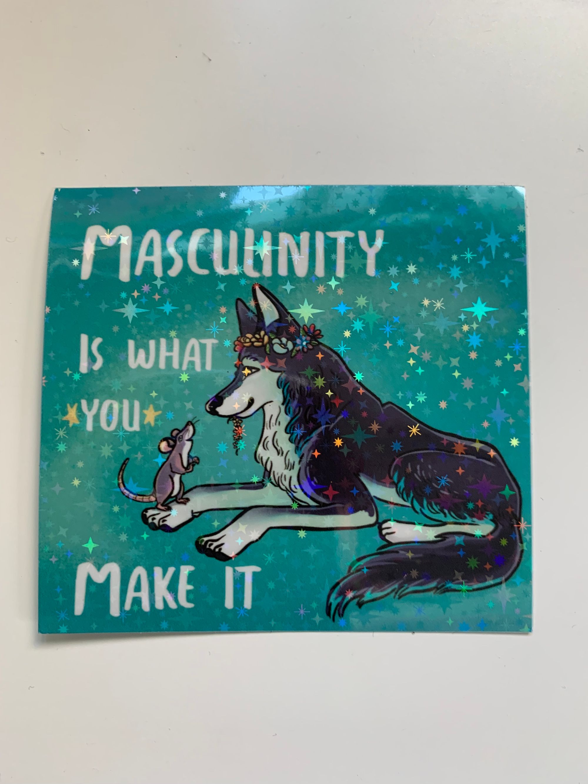 a blue sparkly sticker with a wolf wearing a flower crown & a rat standing on the wolf's paw, being offered flowers. It reads: "Masculinity is What You Make It."