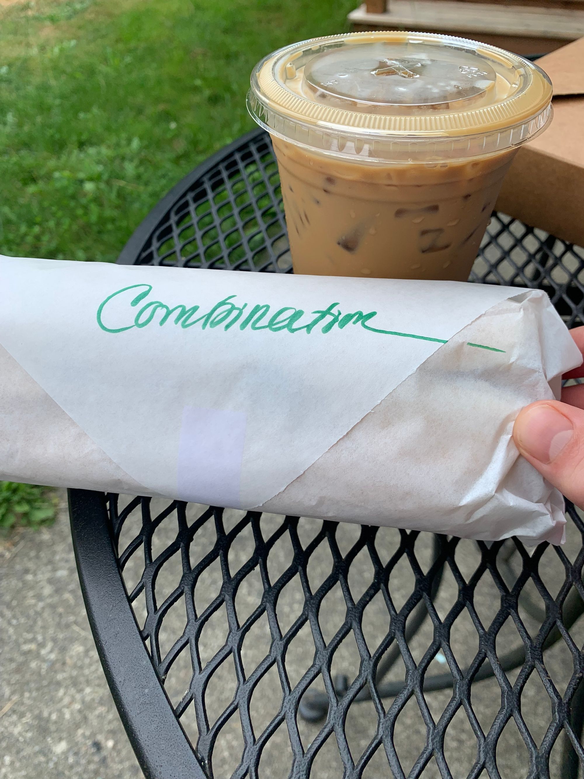 paper wrapped sandwich held by a pink hand on a black metal table, iced coffee and brown takeout box in background