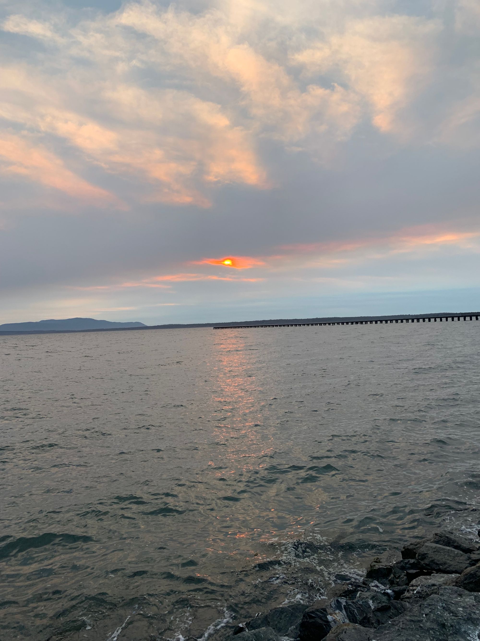 sunset over grey water, long dock in the distance, grey islands on the horizon