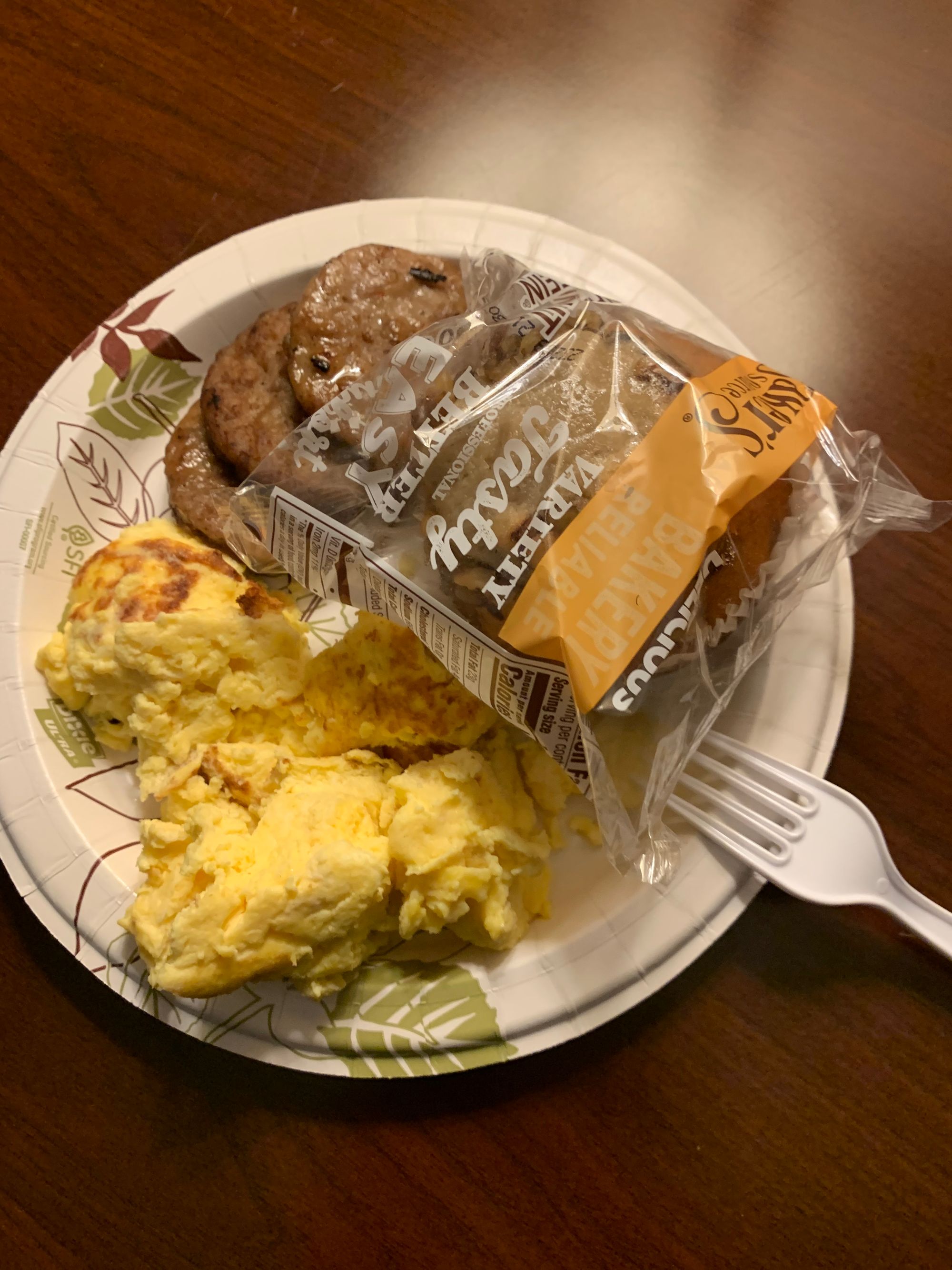 a brown table with a white paper plate holding scrambled eggs, sausage & a packaged muffin, white plastic fork on one side