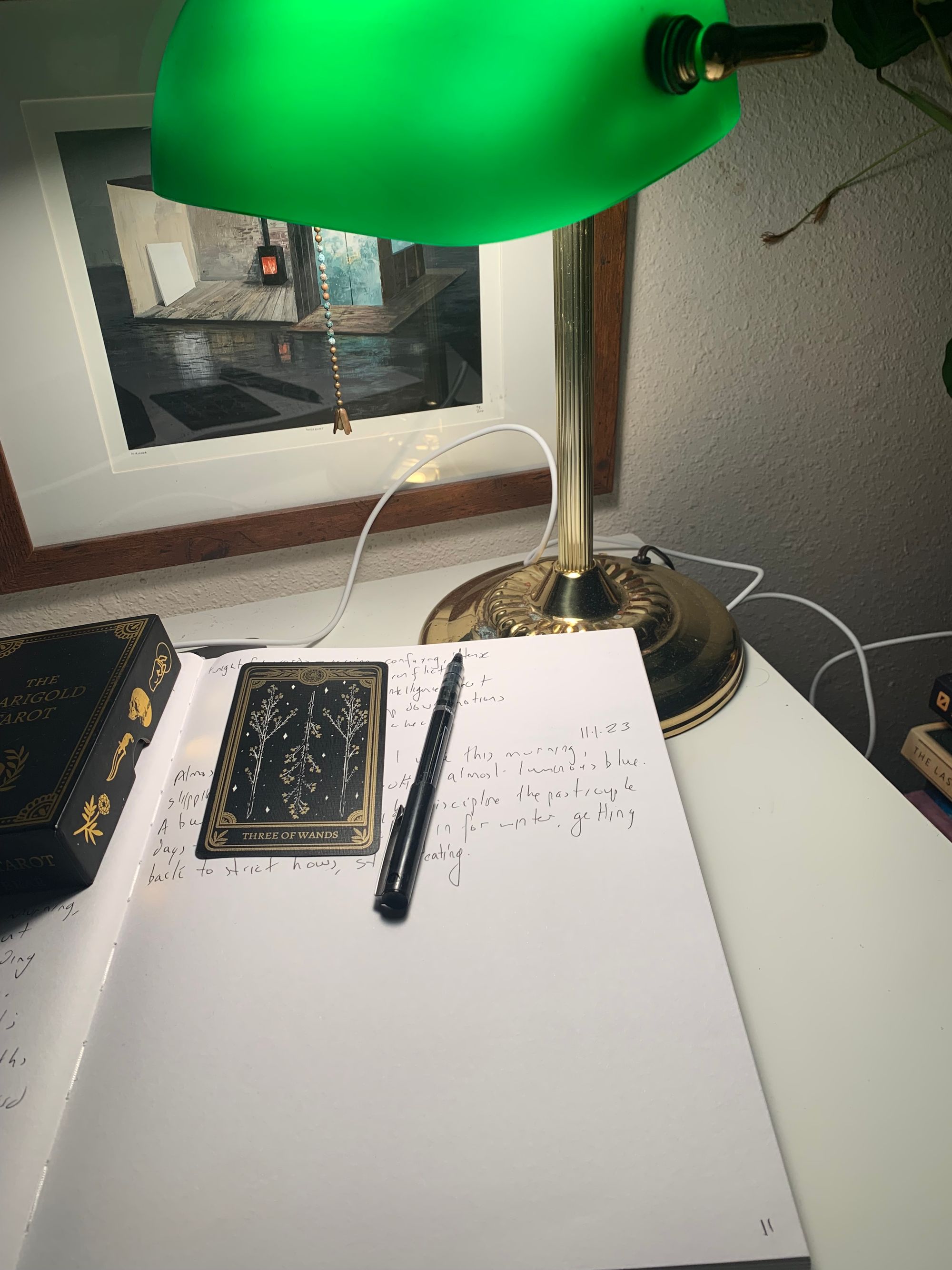 green bankers lamp above a large open notebook, the three of wands tarot car & a pen on a scribbled page