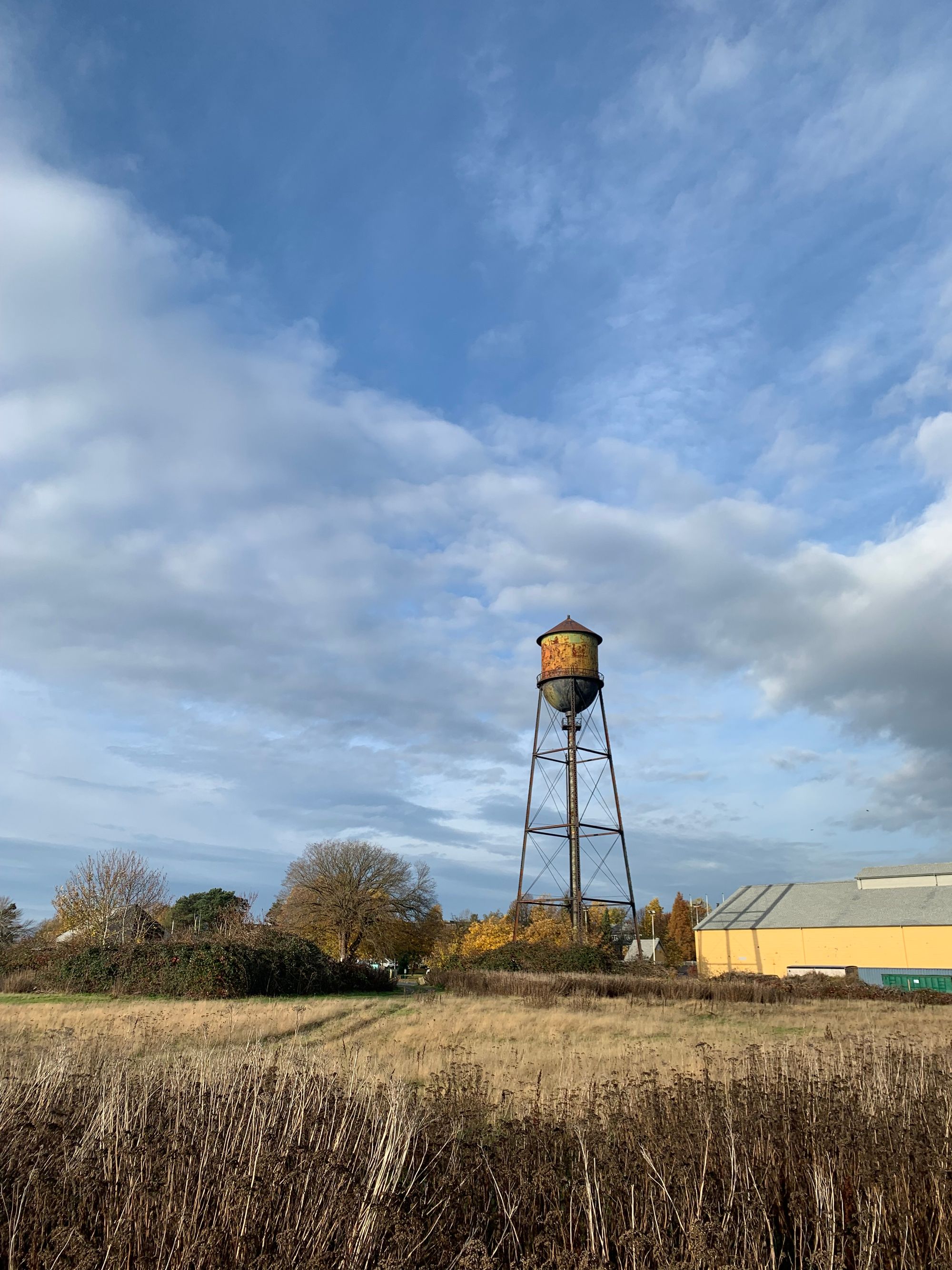 rusty old watertower against blue cloudy sky, dry brown meadow, yellow building on the right