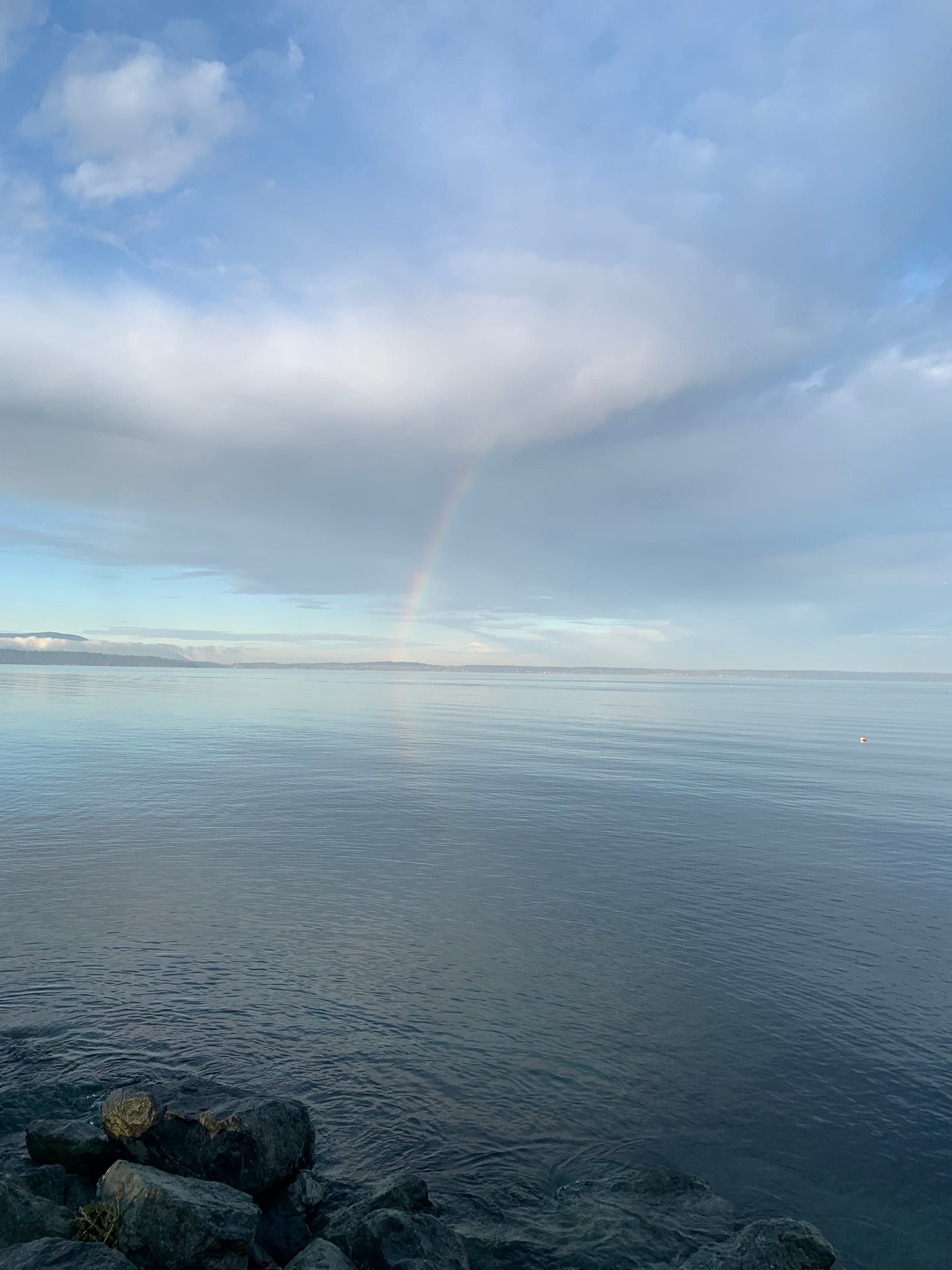 blue cloudy sky, grey blue water, a little partial rainbow touching down on the island across the bay