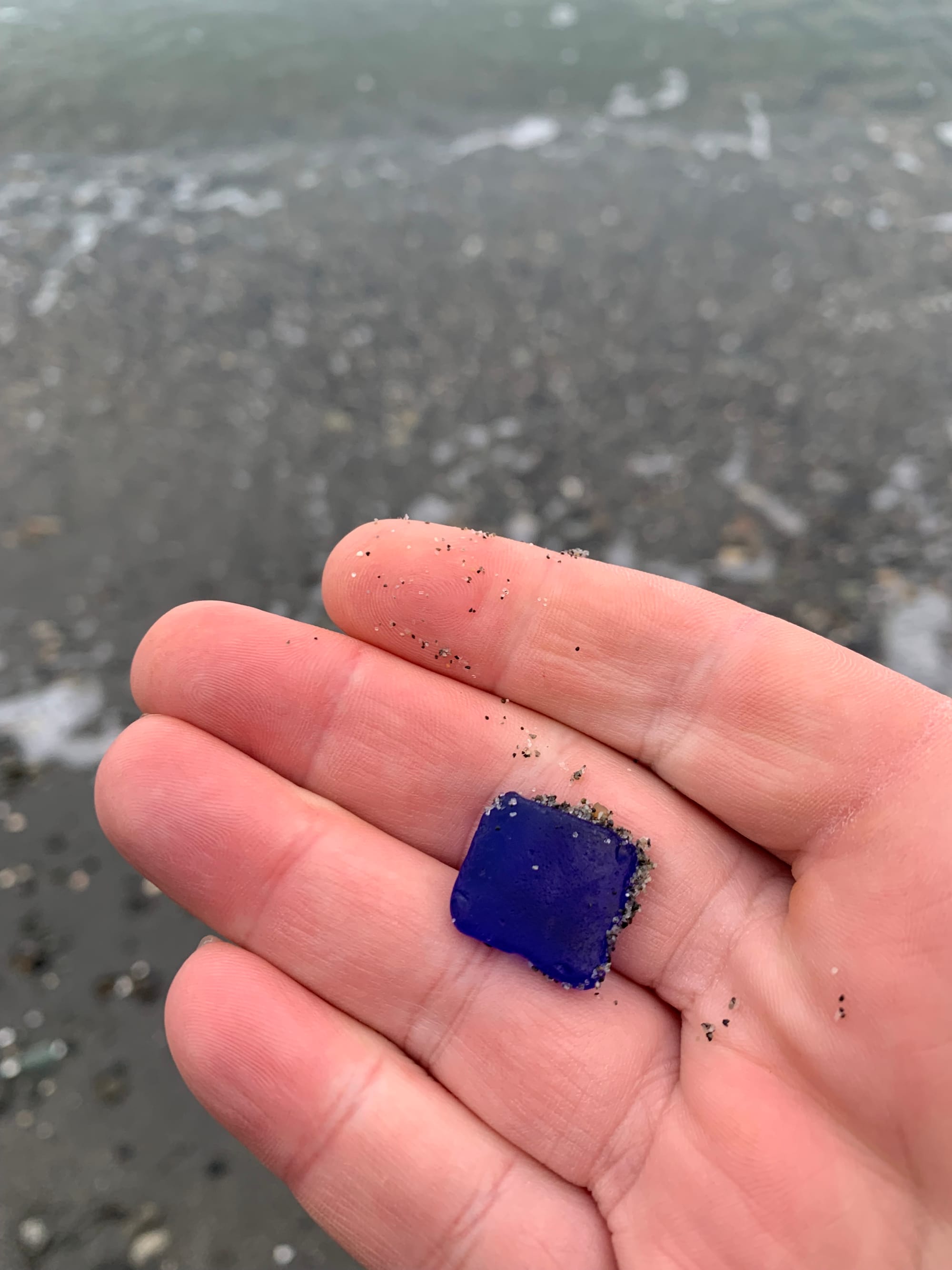 close up of a pinkish hand holding a little square of blue seaglass, background of a pebble beach