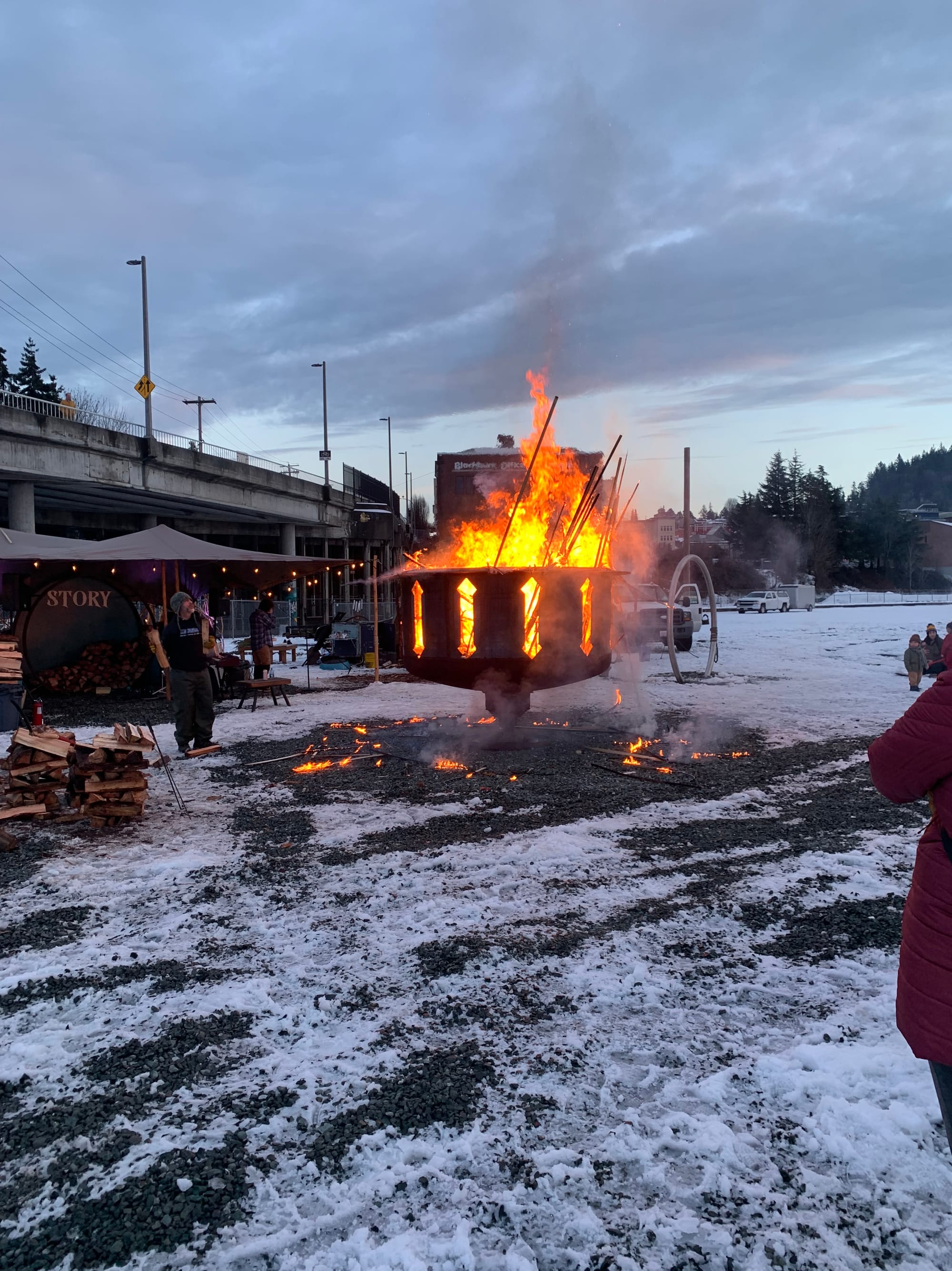 giant metal spinning firepit on a snowy field, guy in black about to toss in some wood, tent behind the firepit with a sign that says STORY