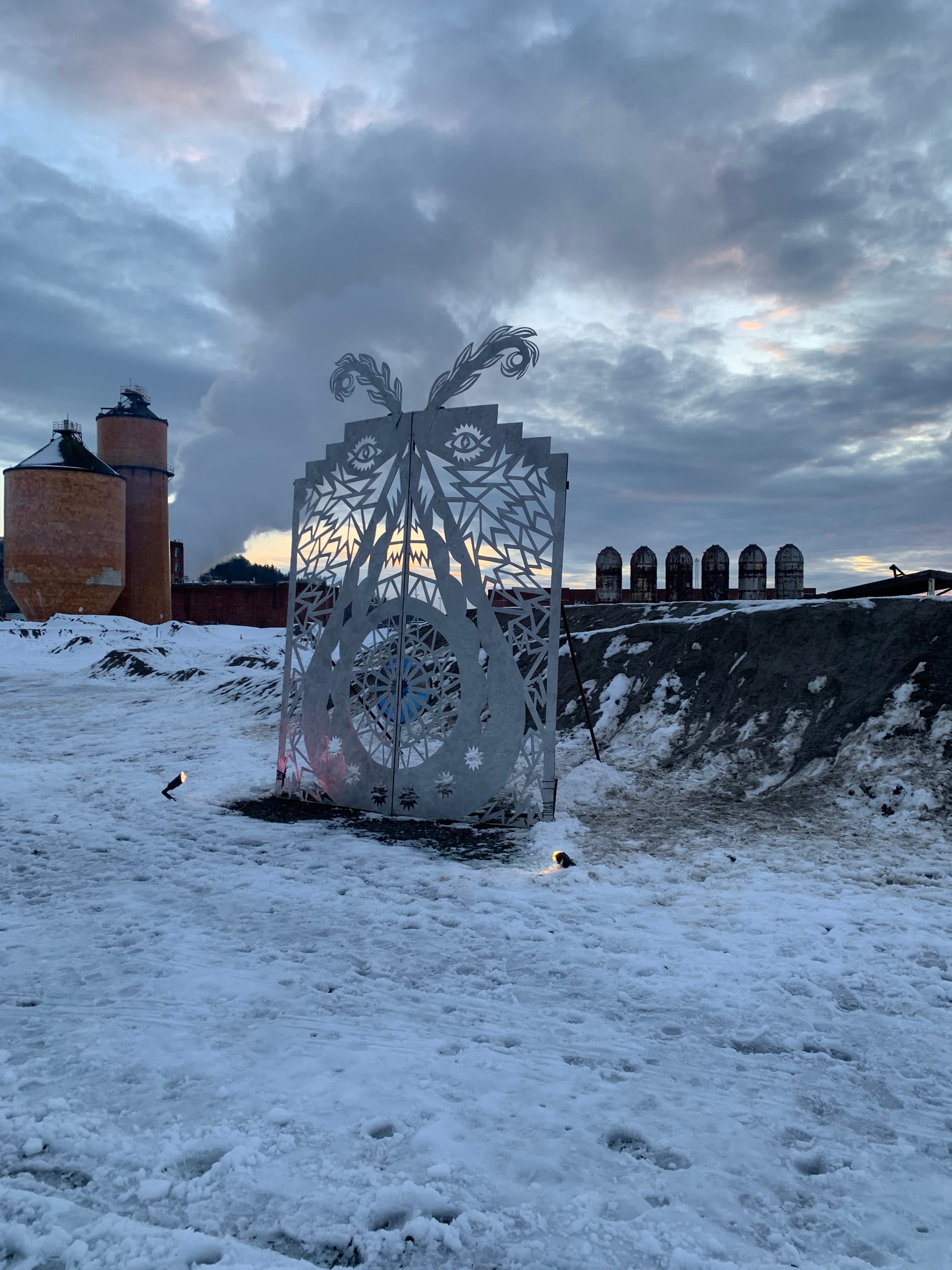 silver metal door art, with feathery antenna & eyes, on a field of snow, industrial equipment visible in the distance