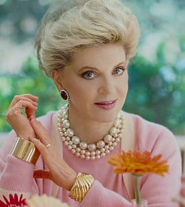 close up of a middle aged white woman with very tall blonde hair wearing a pink cardigan and lot of jewelry, a couple of bright flowers in the foreground