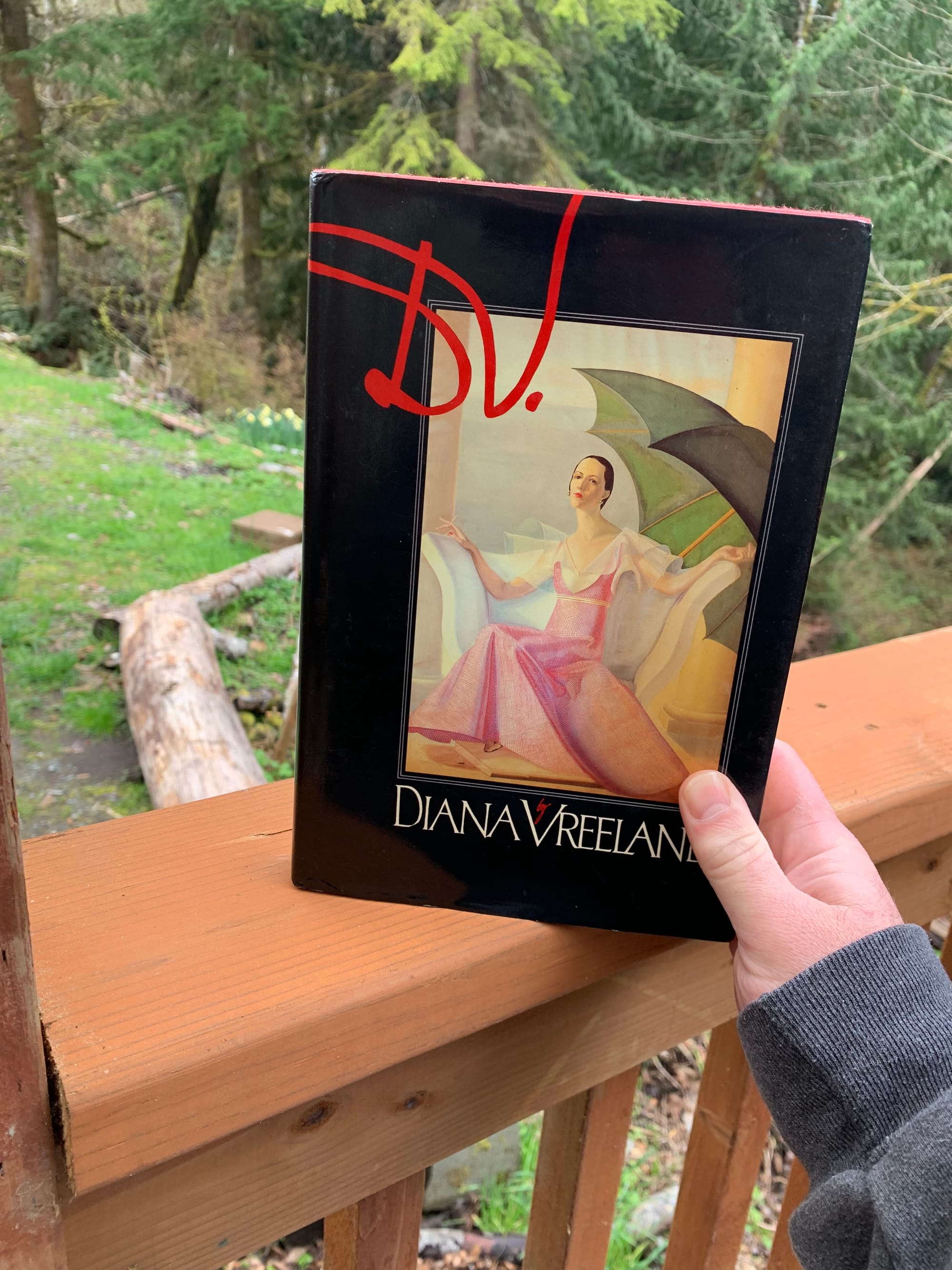 hardback copy of Diana Vreeland's book D.V. held up against a background of a porch railing & trees