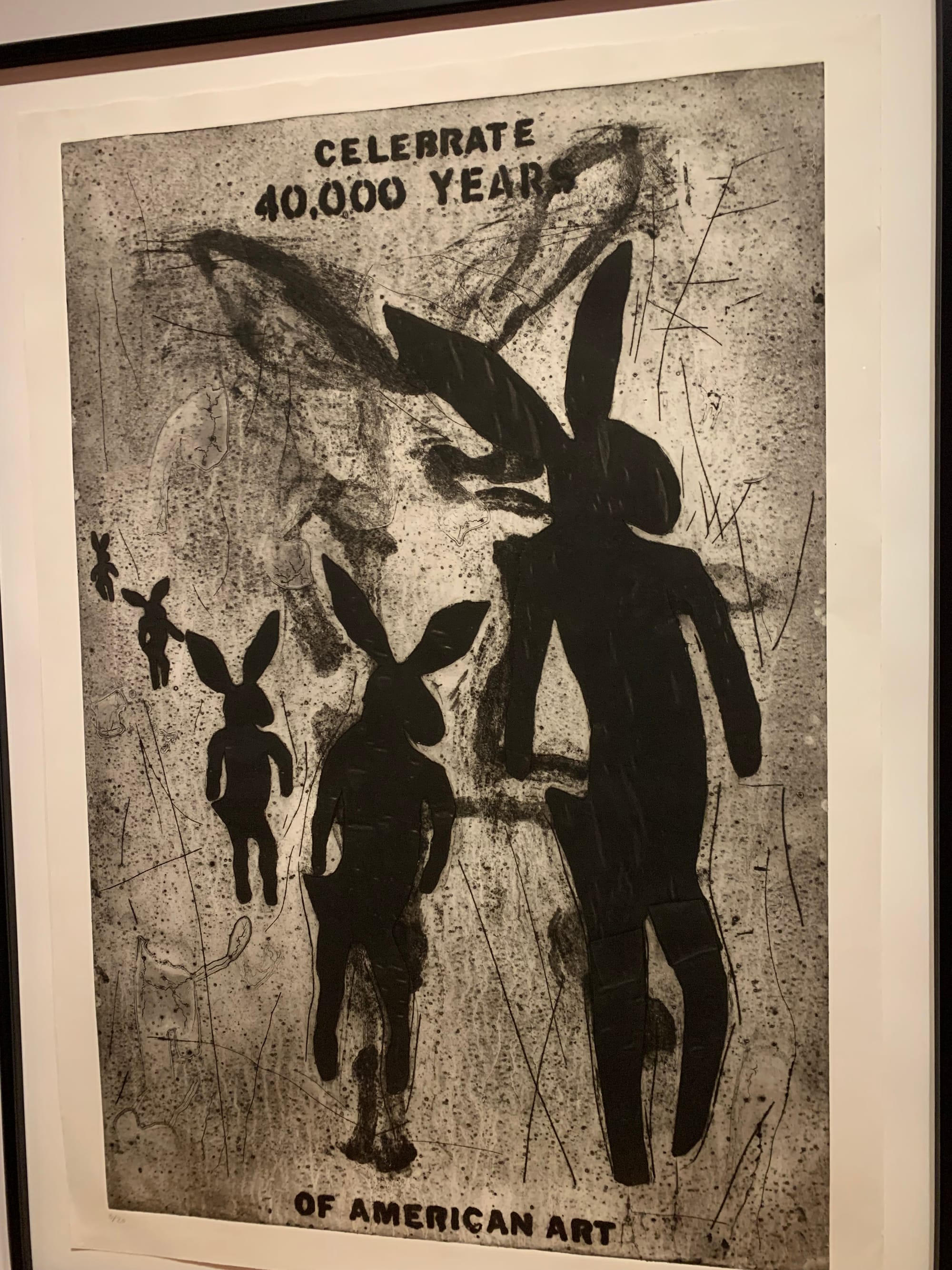 a slightly off kilter photo of Celebrate 40,000 Years of American Art, by Jaune Quick-to-See Smith: a large vertical collagraph etching showing the title & black figures of an upright rabbit