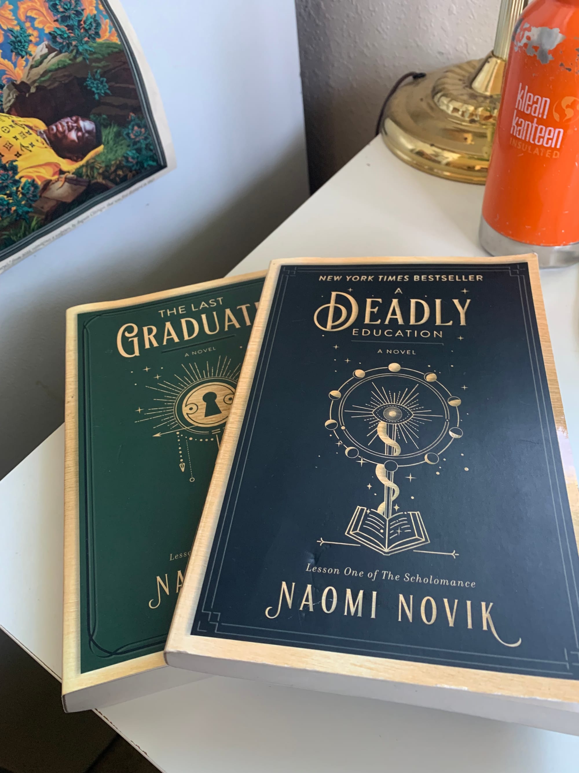 copies of Naomi Novik's A Deadly Education & The Last Graduate, sitting on a white table, water bottle, lamp & brightly colored clipping visible in background