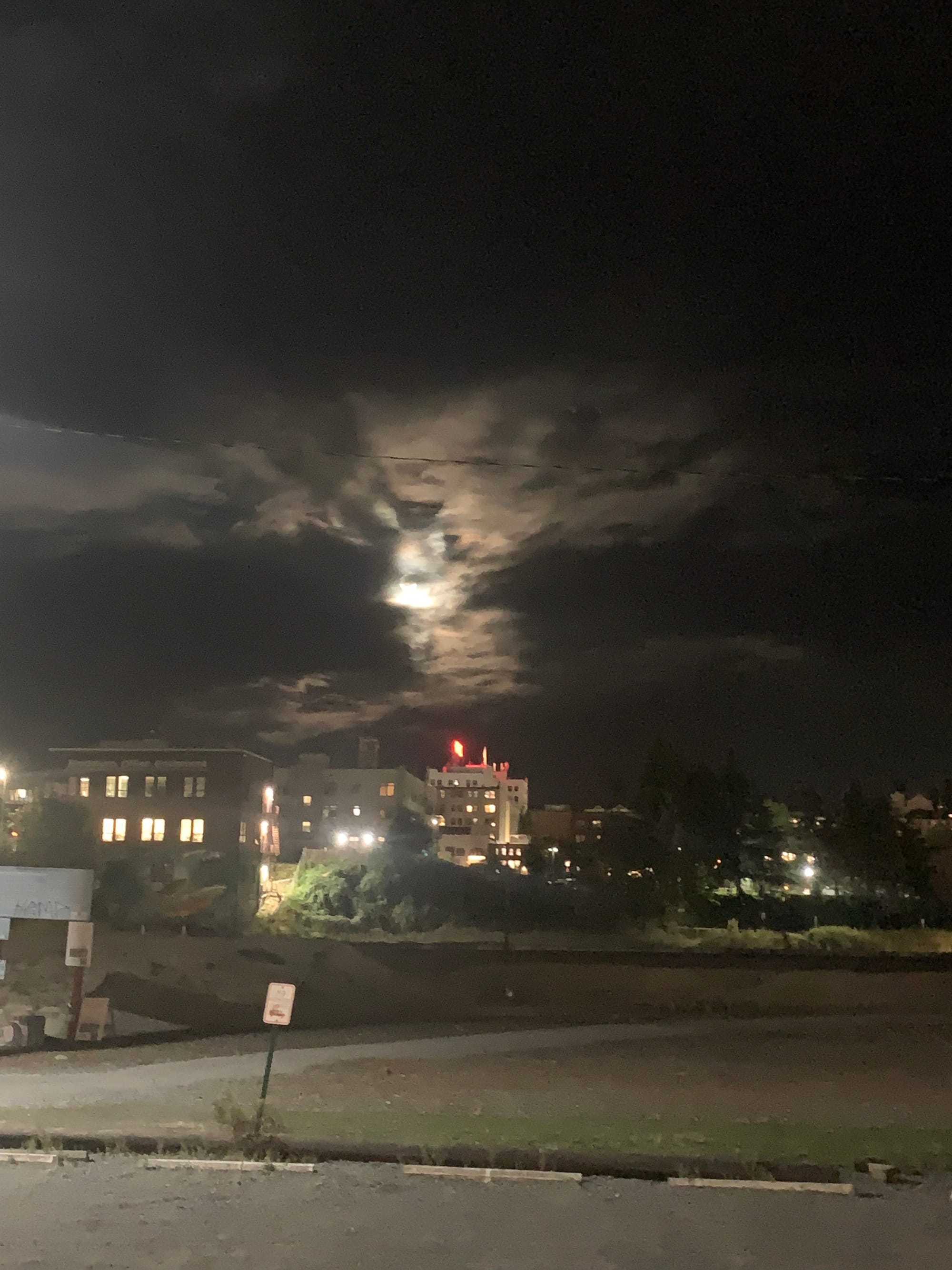 night sky with supermoon visible behind grey clouds, city buildings in the distance, red illegible sign on top of building, empty gravel parking lot