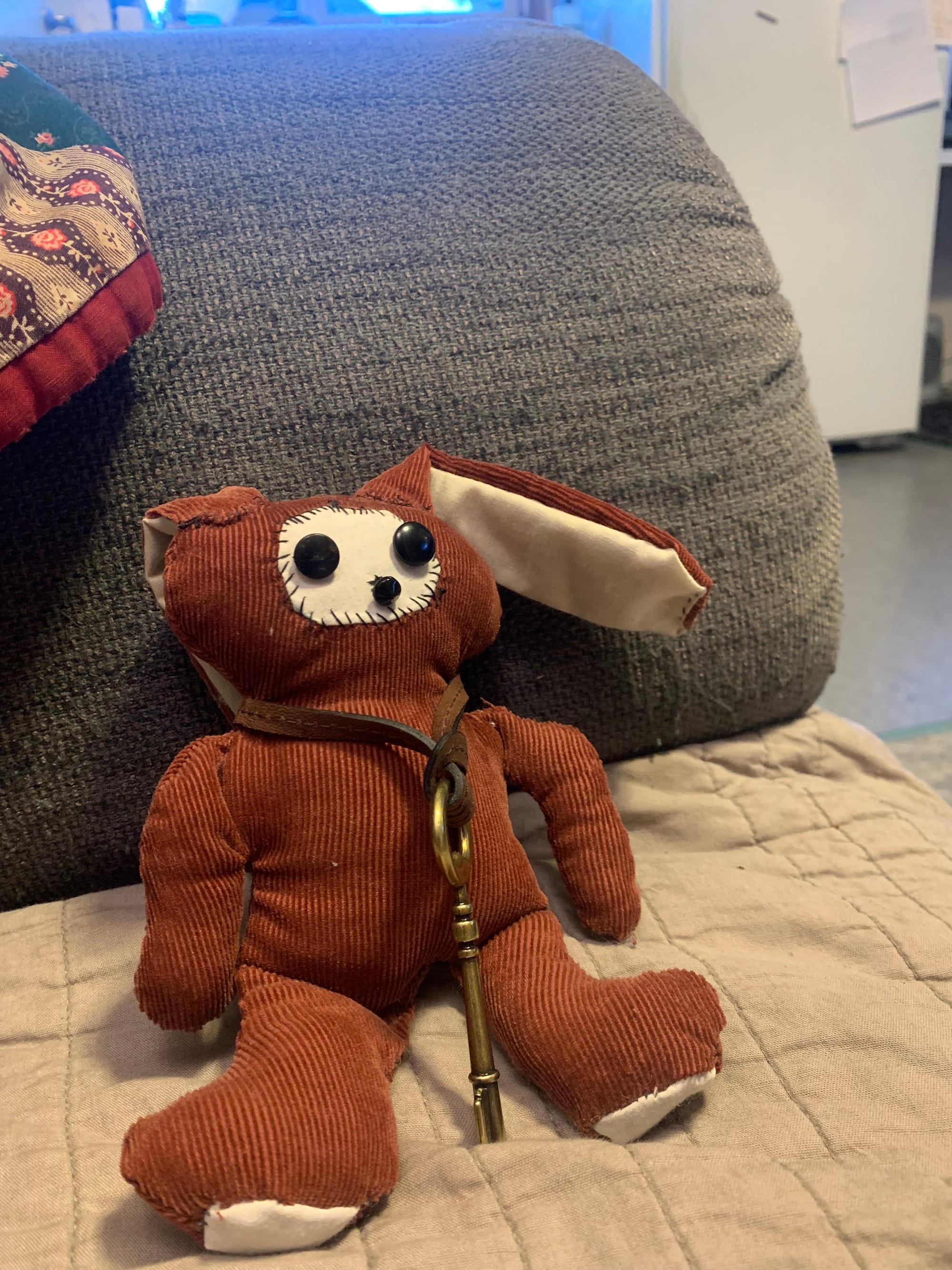 small handmade red corduroy bunny with white paw paws, a white round face & button eyes & nose. a brass key is handing around its neck on a leather strap. 